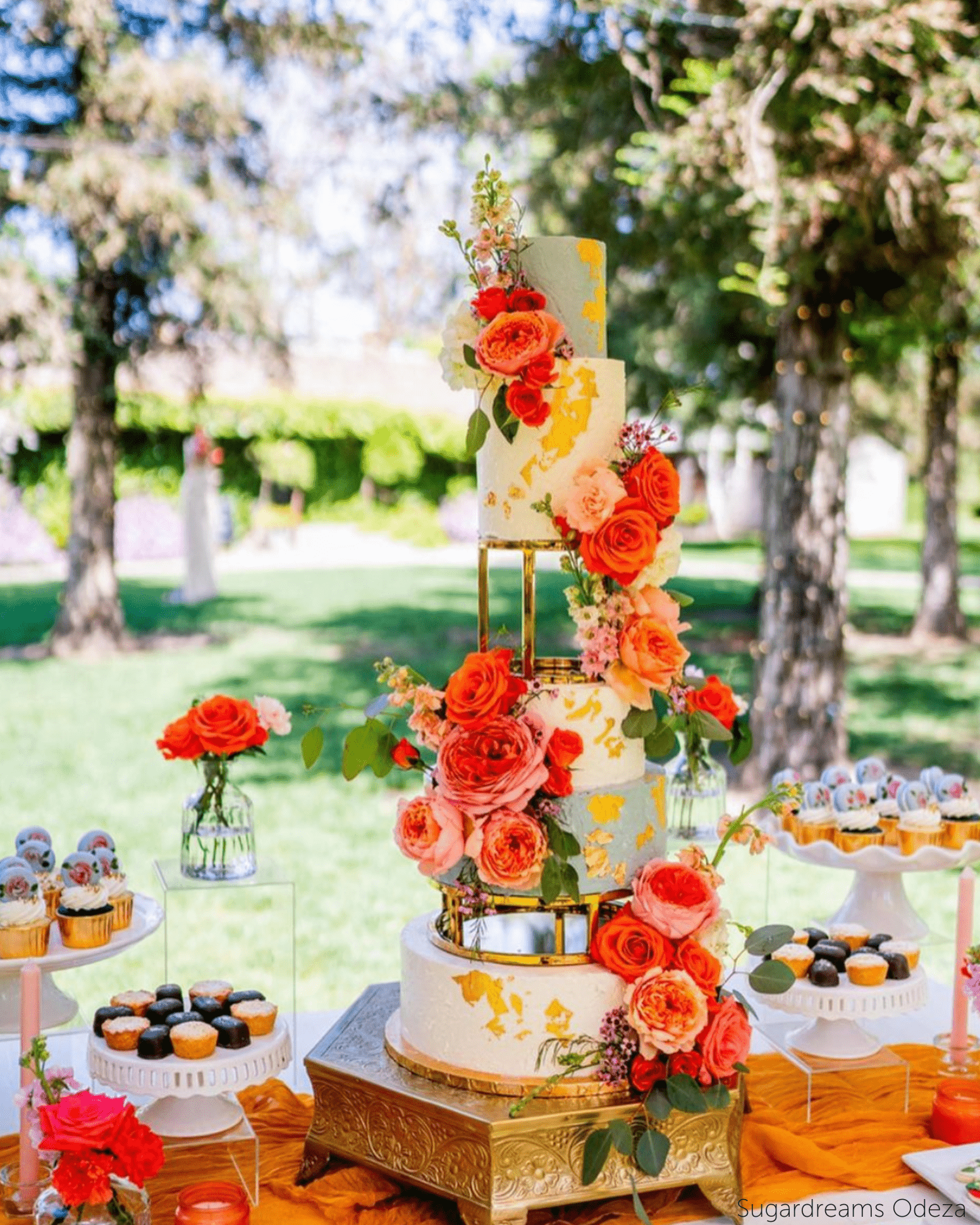 Four-tier outdoor wedding cake with gold metallic round cake separators, bold orange and coral roses, and gold leaf detail displayed on a gold cake stand at an outdoor dessert table.