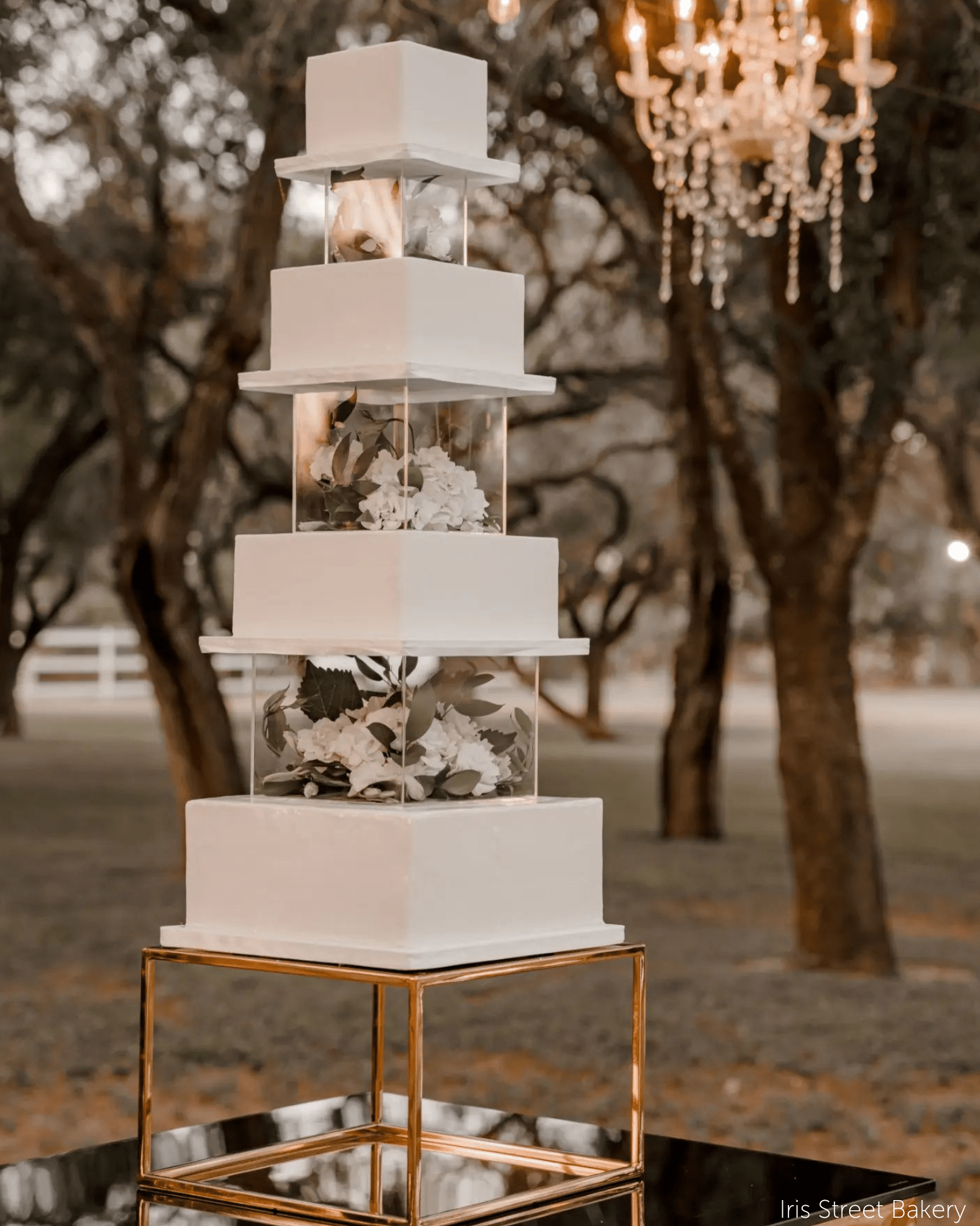 Four-tier square wedding cake with clear acrylic fillable tiers and white florals displayed on a metallic gold cake plinth in an outdoor evening setting.