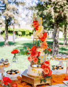 Four-tier outdoor wedding cake with gold metallic round cake separators, bold orange and coral roses, and gold leaf detail displayed on a gold cake stand at an outdoor dessert table.